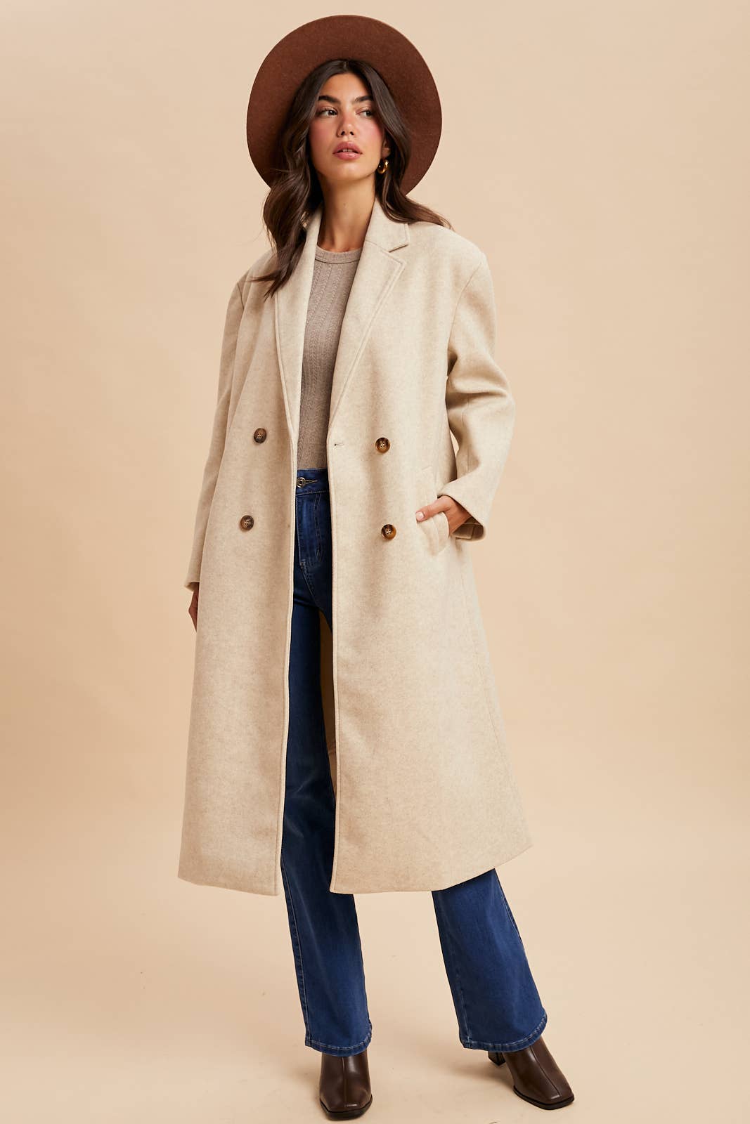 Woman wearing a beige coat, brown hat, and blue jeans on a beige background